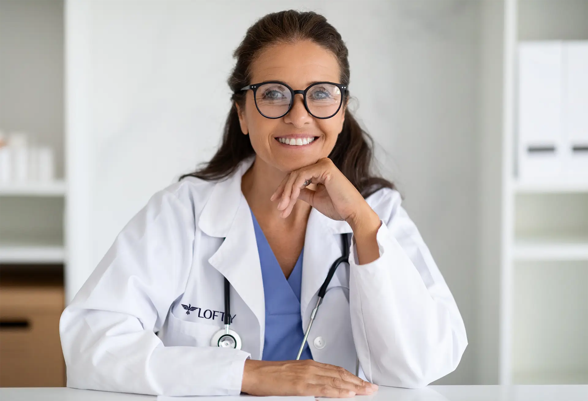 A doctor with long brown hair and glasses smiles while sitting at a desk. She is wearing a white coat and has a stethoscope around her neck. The background is a tidy office setting.