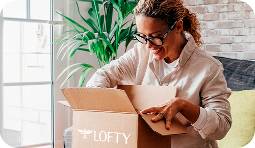 A person with curly hair and glasses is sitting on a couch, joyfully opening a cardboard box labeled "LOFTY." The room is bright, with a large plant and a brick wall in the background.